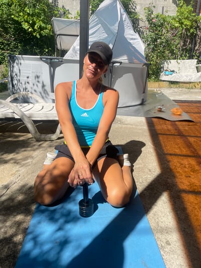 A woman is kneeling on a blue yoga mat outdoors, holding a dumbbell. She is wearing a black baseball cap, a blue tank top with an Adidas logo, and black shorts. She has blonde hair and is smiling at the camera. The background shows a backyard scene with a lounge chair, a large grey inflatable pool, and lush green vegetation. The ground is a mix of concrete and reddish-brown stained concrete. Sunlight casts strong shadows across the scene. The location is identified as Baranovka, Russia.