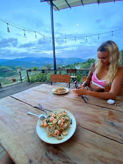 An outdoor restaurant with a view of a valley at dusk. A woman with blonde hair and tattoos sits at a wooden table, looking at her phone. In front of her are two plates of food, chopsticks, and a spoon. String lights hang above the table, and a partially visible roof is overhead. The background shows rolling hills with some buildings and vegetation. The sky is cloudy, with a sliver of the moon visible. The restaurant appears to be located in Thôn 3, Vietnam, given the context.