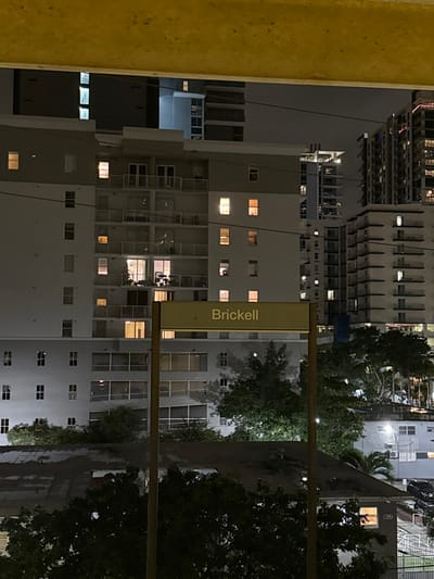 A night cityscape features the "Brickell" station sign in the foreground. Buildings of varying heights surround the station, some with illuminated windows. Trees dot the scene, casting shadows and adding greenery to the urban environment. The overall atmosphere is dark, lit by artificial lights. The image is rotated, requiring the viewer to adjust their perspective.