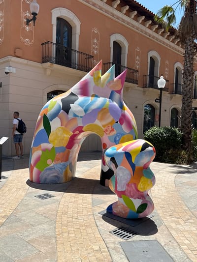 A man wearing a white t-shirt and denim shorts with a black backpack stands looking to his left in front of a brightly colored abstract sculpture. The sculpture is painted with various pastel and vibrant abstract shapes and colors. Behind the sculpture, a building with an orange facade features arched windows with black balconies and decorative white trim. A palm tree is visible to the right of the building. The ground is paved with light-colored square and rectangular stones. The scene appears to be outdoors in a public area, possibly a shopping district or plaza. The location is identified as La Roca Village, La Roca del Vallès, Spain.