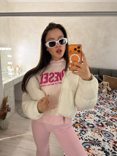 A young woman stands indoors, facing a mirror, taking a selfie with an orange-cased phone. She wears white sunglasses, a white puffer jacket over a pink top with a reversed word in magenta print, and pink leggings. Her tongue is slightly visible. She is holding up two fingers. Behind her, a vanity table with illuminated mirrors is visible. The background includes a patterned bedspread and a glimpse of a gray couch. It's not possible to determine if the image was taken in Pernik, Bulgaria.