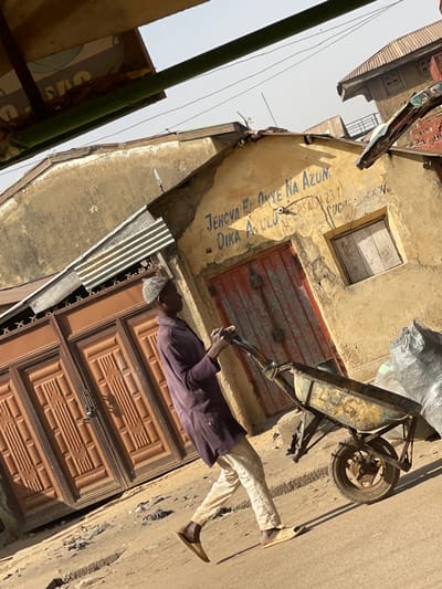 In Zaria, Nigeria, an outdoor scene depicts a male subject walking from left to right, pushing a metal wheelbarrow containing a grey bag. The man wears a light-colored cap, a dark purple long-sleeved tunic, light-colored trousers, and light brown slip-on sandals. He is positioned on an uneven, unpaved surface under direct sunlight, indicated by strong shadows.
Behind the subject, a series of weathered buildings are visible. The central building features light tan or yellowish stucco walls with visible peeling and cracks. Its facade prominently displays faded blue text: "JEHOVA BU ONYE NA AZUN", "DIKA A.ULU", and "PSALM 23:1". This building has a distressed, double-leaf, red-painted wooden door with vertical panels and a small, boarded-up window to its right. To the left of the central building, a large set of paneled brown double gates, secured with a padlock, is partially visible. Corrugated metal sheets are affixed above these gates, serving as a rudimentary awning or roofing extension. Additional building structures with corrugated metal roofs are discernible in the background. The sky is hazy light blue, and overhead, thin wires and a partial view of a green structural element are present in the upper frame. The entire image has a slight diagonal tilt.