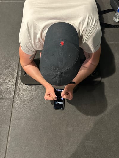 The image shows a person doing a plank exercise. The person is wearing a black baseball cap with a red logo and a light-colored t-shirt. Their forearms are resting on the floor, and their body is in a straight line from head to toe. A smartphone is placed on the floor in front of the person, displaying a timer that reads "00:34.55". The person's hands are clenched into fists. The exercise appears to be taking place on a dark-colored tiled floor, possibly in a gym or a home workout space. No specific location in Alès, France, can be determined from the visual information.