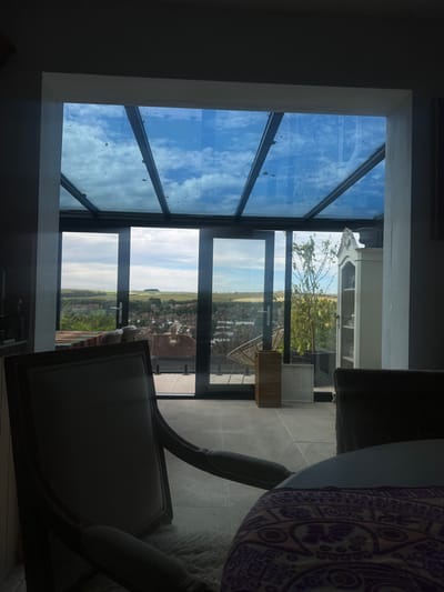 This image captures an interior view looking out through a large, multi-paned glass ceiling and windows. The scene is a room with a dark wall on the left, and a large table partially covered with a patterned cloth on the right. A chair with a light-colored seat is in the foreground, also on the right. Beyond the glass, a landscape unfolds under a partly cloudy sky. The view appears to be of a town or village with houses, fields, and trees, suggesting a rural or semi-rural setting. A thin tree with some green leaves and possibly fruit is visible closer to the window. The lighting suggests it is daytime, with a mix of bright sky and shadows within the room, indicating possibly late morning or afternoon. The overall impression is one of a comfortable indoor space with an expansive view of the outdoors. There is no visible text in the image.