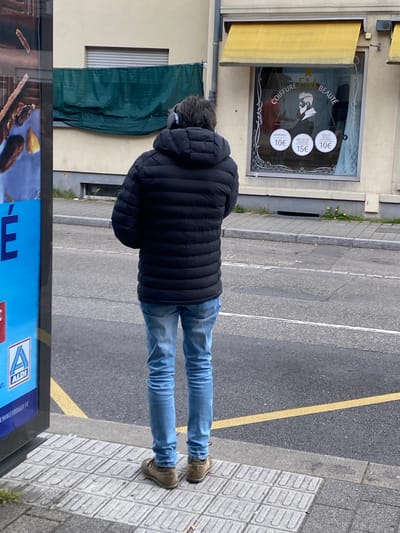 The image shows a person in a black puffy jacket and light blue jeans standing on a paved sidewalk in Bischheim, France. They are wearing headphones. A yellow line divides the sidewalk from the asphalt road. Next to the person is a bus stop advertising board that displays a food advertisement and the ALDI logo. Behind the person is a building with a hair salon ("COIFFURE LM BEAUTÉ") sign in the window, listing prices for services such as "DEFRISAGE" and "MECHE DE CHEVEUX" and "SOIN DE VISAGE EPILATION". A green tarp is partially covering a window to the left of the salon.