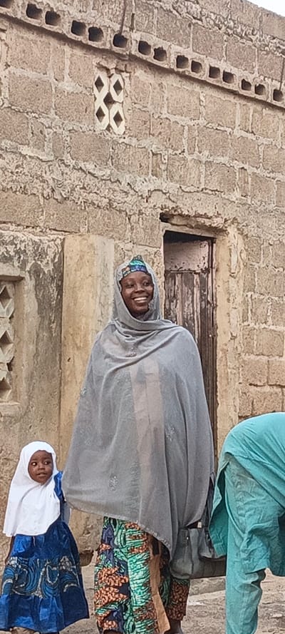 This outdoor portrait captures a vibrant street scene in Rangaza, Nigeria, featuring a smiling woman, a child, and a partially visible third person against a rustic stone building.
In the center, a woman grins broadly, her face radiating joy. She wears a grey head covering over a colorful, patterned cap, a long grey outer garment, and a brightly patterned wrapper or skirt in hues of green, blue, and orange. A dark shoulder bag hangs from her left side. To her left, a young child, likely a girl, stands wearing a white head covering and a blue and white patterned dress. The child gazes to her left with a neutral, inquisitive expression. On the far right, another person is partially visible, dressed in a light blue or turquoise garment, appearing to be bent over or obscured.
The background is dominated by a sturdy, light-colored stone wall constructed from rough-hewn blocks. Decorative elements are embedded in the wall, including a white geometric pattern resembling connected diamond shapes and a horizontal band of small square openings. A dark, aged wooden door is visible behind the main woman. The bright, even illumination suggests it is daytime. The scene conveys a sense of community and daily life, with the main subject's happy expression being a notable detail. No visible text is present.