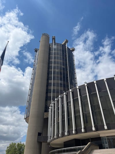 The image shows a low-angle shot of the Palais de Justice in Créteil, France, under a bright, cloudy sky. The building features a prominent, cylindrical concrete tower with vertical ribbed detailing. Adjacent to the tower is a glass-and-steel structure with a dark, reflective facade. The building's architectural style appears to be modernist. A French flag on a flagpole is visible on the left side of the frame. The environment suggests an outdoor urban setting on a daytime. There are no people visible in the image.