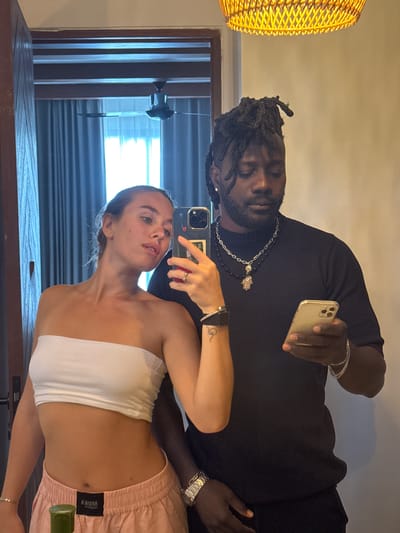 The image shows a young couple taking a mirror selfie in what appears to be a hotel room. The woman, with blonde hair pulled back, is wearing a white bandeau top and pink athletic shorts. She is holding a smartphone in her right hand, which she is using to take the photo. A silver ring is visible on her left hand. She has a small tattoo of a snake on her left forearm.
The man, with dark skin and dreadlocks, is wearing a black t-shirt and multiple silver necklaces. He is holding a smartphone in his right hand and looking at it. A silver bracelet is visible on his left wrist.
The background reveals a room with beige walls, a wooden door frame on the left, and dark blue curtains covering a window behind the couple. A ceiling fan is visible in the upper left corner of the frame, and a woven light fixture hangs from the ceiling. The overall impression is that of a casual, indoor setting, likely a hotel room.
This image is not in Kediri, Indonesia.
