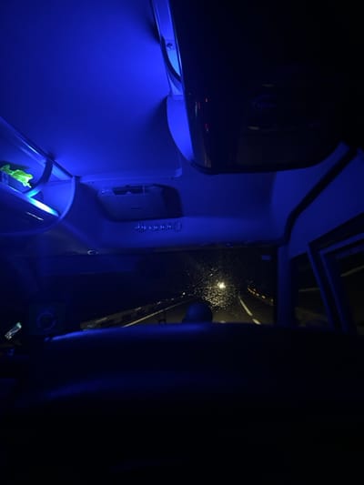 The image shows the interior of a vehicle traveling at night. The driver is visible from the back, with a dark silhouette. There is a bright blue light emanating from the ceiling of the vehicle and the driver's side window is slightly open, showing a view of a highway lit by rain-soaked streetlights. The vehicle is traveling on a highway in La Losa, Spain at coordinates 40.842, -4.149.