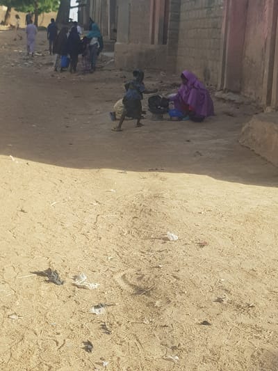 The image depicts a dirt road in what appears to be a residential area, likely in Dabawa, Nigeria. Several individuals, possibly women, are gathered along the side of the road. Some appear to be carrying items on their heads or working on the ground. The ground is covered in sand and debris. The background shows buildings with weathered walls. The lighting suggests it is daytime.