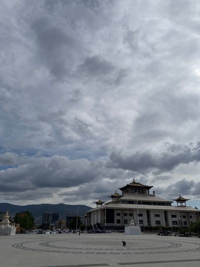 A building with a multi-tiered roof stands adjacent to a gray paved plaza, with outlines of circular designs, located in Ulaanbaatar, Mongolia. Mountains form a backdrop. Clouds dominate the sky. Cars are parked between the building and the mountain. A person is visible in the plaza.