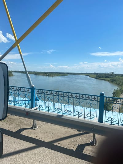 The image captures a view from a bridge overlooking a wide, tranquil river under a bright blue sky with scattered white clouds. The foreground shows a section of the bridge's blue decorative railing and a metal guardrail. Two yellow-wrapped poles, possibly part of the bridge's infrastructure or an art installation, cross diagonally in the upper left portion of the frame. The river flows from the left, curves to the right in the distance, and is bordered by lush green trees and vegetation on both banks. The distant landscape features scattered buildings and power lines, suggesting an urban or semi-urban environment. The overall scene is peaceful and serene, bathed in natural daylight. The location of Aqsu city administration, Kazakhstan, is not explicitly evident from the visual elements in the image itself but is provided as contextual information.