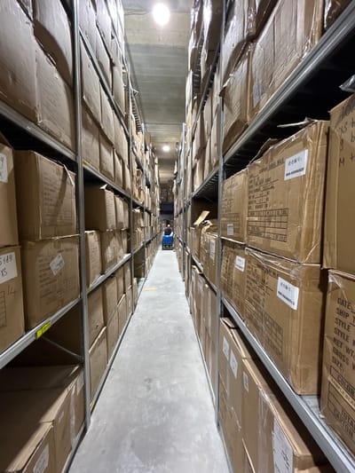 This image shows a warehouse aisle filled with tall shelves stacked with cardboard boxes. The shelves are made of metal and are organized in a grid pattern. The boxes are mostly brown and appear to be uniform in size, suggesting they contain similar items. Some boxes have labels with text and numbers, indicating inventory information. The aisle is well-lit by overhead lighting. In the distance, a person is visible pushing a blue cart down the aisle, suggesting that the warehouse is operational. The concrete floor is clean and shows some signs of wear. The overall environment is that of a storage facility. Given the context of Holon, Israel, this could be a distribution center, a logistics hub, or a storage facility for a company operating in the region.