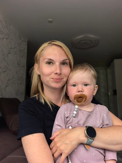 A blonde woman in a black t-shirt holds a baby wearing a pink onesie with a pacifier. The woman has yellow eyeliner and a watch on her left wrist. The baby has blue eyes and chubby cheeks. They are in an indoor setting with a patterned wall and a dark ceiling. The background suggests a living room or apartment, with a glimpse of a couch. The image is a portrait-style photo, focusing on the two subjects. The specific location within Sterlitamak, Russia, cannot be determined from the visual information provided.