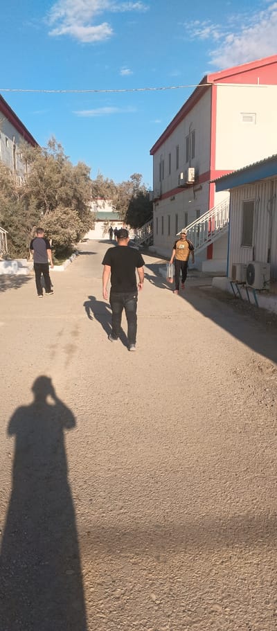 The image depicts a sunny outdoor scene in Balkanabat City, Turkmenistan. In the foreground, a person's shadow is cast on a gravelly path. Further down the path, three men are walking away from the camera. The man closest to the camera is wearing a black t-shirt and dark jeans. To his left, a man in a blue t-shirt and black pants walks ahead. To the right, a man in a yellow and black t-shirt and black pants carries a plastic bag. Two buildings, one with white and red accents and another with grey siding, flank the path. Trees and vegetation are visible between the buildings and in the background. The sky is blue with scattered white clouds.