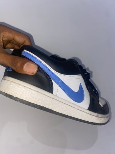 A hand holds a black, white, and light blue Nike Air Jordan 1 Low sneaker. The shoe has a black toe box, white side panels, and a light blue swoosh. The midsole is white with a black outsole. The shoe appears used, with slight creasing and wear on the sole. The background is a light grey or white.