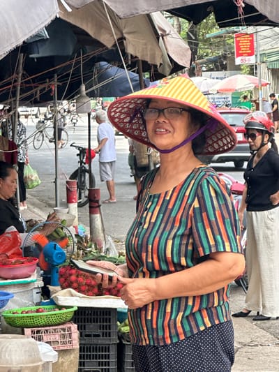 In Mai Chau, Vietnam, a street scene unfolds under a canopy of umbrellas and tarps. In the foreground, a woman wearing a conical hat adorned with yellow and red stripes, glasses, and a colorful striped shirt gazes forward while holding a smartphone. To her right, another woman wearing a red helmet and sunglasses stands with her hands on her hips. A red car is visible in the background. Stalls line the street, displaying various fruits, including rambutans and other red produce, alongside baskets, crates, and an electric fan. Pedestrians, a bicycle, and motorbikes are present, contributing to the bustling atmosphere of the marketplace. The ground is paved with concrete.