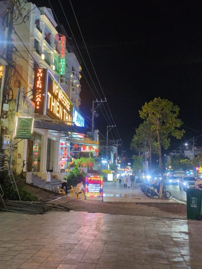 This is a night-time street scene in Phú Quốc, Vietnam. A brightly lit street displays a variety of signs. The most prominent signs advertise "VIP MASSAGE THIEN THA" and "GALAXY HOTEL," along with an illuminated sign for "Miana Resort." Colorful hanging lanterns are visible. The scene features a sidewalk with tiled paving. A parked motorbike and a trash receptacle are at the edge of the sidewalk. People are visible in the distance walking along the street. Numerous electrical wires are overhead.
