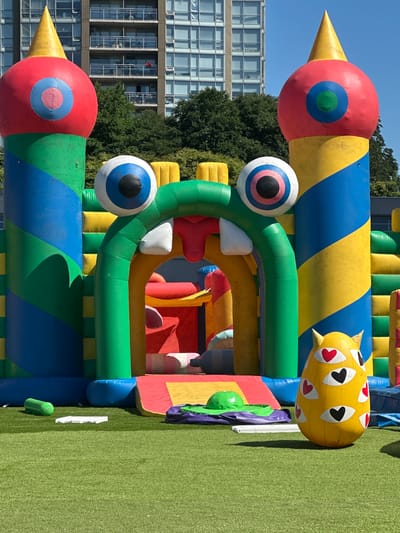 The image displays a vibrant, inflatable playground structure set against a backdrop of a multi-story building and trees under a clear blue sky. The playground is predominantly colored in red, yellow, green, and blue, with large, exaggerated features such as eyes and conical towers. In the foreground, a bright yellow inflatable object with multiple heart-shaped eyes is positioned on a green artificial turf surface. Several smaller inflatable elements and a green mat are also visible on the turf. The building in the background appears to be an apartment or office complex with numerous windows. The overall scene suggests an outdoor recreational area, likely a park or a community event space, in the city of Coquitlam, Canada. No people are visible in the image.
