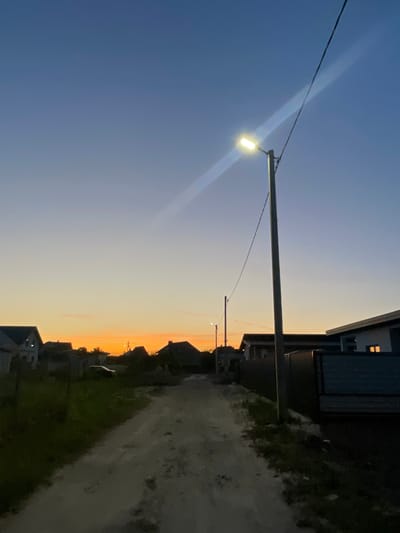 The image depicts a rural scene in Belarus at dusk. A single streetlamp illuminates a dirt road lined with houses and vegetation. The sky transitions from a deep blue to a soft orange as the sun sets, casting a warm glow over the landscape. The overall mood is peaceful and serene, evoking a sense of quietude and rural charm. There is no visible text in the image.