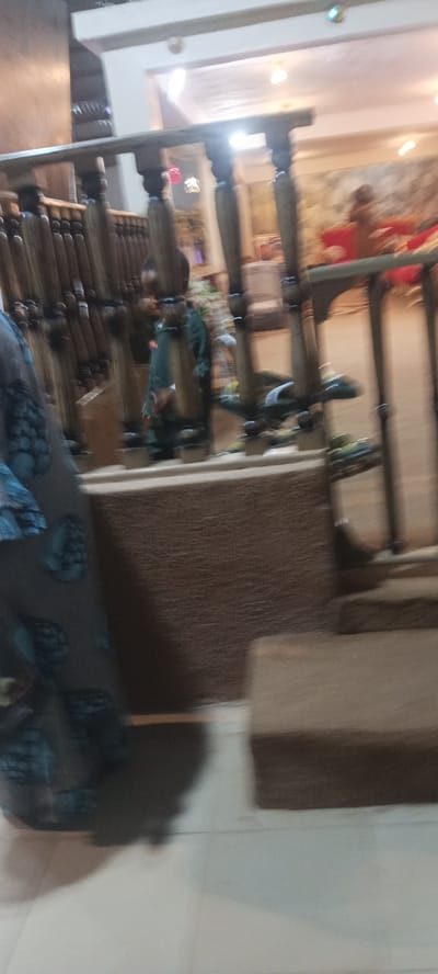 The image depicts a blurred interior scene, possibly a staircase or landing area with wooden railings. A person wearing a patterned garment is partially visible on the left side of the frame. In the background, a person in a dark green outfit appears to be walking or standing near the railing. The environment features wooden architectural elements, a textured wall, and some faint lighting from above. The overall impression is of a dimly lit, indoor space with some decorative elements. There is no visible information in the image to confirm its location in Rukuba Road, Nigeria.