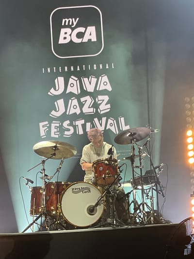 The image depicts a stage during the Java Jazz Festival in Salembaran, Indonesia. A drum set is positioned center-right, with cymbals and a snare drum visible. A drummer with short, light-colored hair is seated behind the drum set. Behind the drummer and the drum set is a large screen displaying the event logo: "my BCA International JAVA JAZZ FESTIVAL." The lighting suggests an indoor venue with stage lights visible at the top of the frame. The scene conveys an active concert environment.