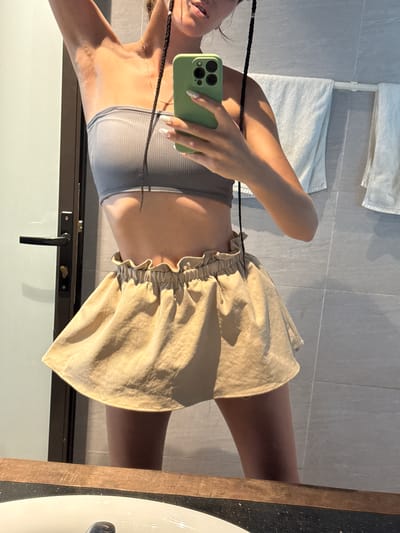 A mirror selfie captures a person's upper body and legs, rotated ninety degrees counter-clockwise. The individual, likely a woman, is seen from the chest down to the mid-thighs, holding a light green smartphone with a distinct camera array (resembling an iPhone) to take the picture. She wears a light grey ribbed crop top that exposes her midriff and a light beige or tan mini skirt with a ruffled elastic waistband. A thin gold chain necklace is visible around her neck, and long, dark braided hair hangs over her shoulder, extending into the shot. Her fingernails are painted with a light, possibly iridescent, polish.
The setting appears to be a brightly lit bathroom, indicated by the light grey tiled walls and two white towels hanging on a rack in the background. Strong natural light, possibly sunlight, comes from the left, casting shadows on her arm, neck, and the folds of her skirt. To the right, part of a wooden-edged countertop and a white sink basin are visible. A dark door handle is also present in the lower right portion of the frame. The overall impression is a casual indoor scene. There is no visible text in the image. Given the provided context, the location is Hà Giang 1 Ward, Vietnam.