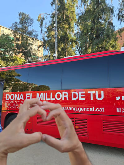 In an outdoor setting, likely in the city of Cornellà de Llobregat, Spain, a red bus with white lettering is prominently featured. The text on the bus reads "DONA EL MILLOR DE TU" and below it "DONA" and "arsang.gencat.cat". The bus has large tinted windows. In the foreground, two hands are forming a heart shape, partially obscuring the lower portion of the bus. The background shows trees and buildings, with a clear blue sky overhead. There are also streetlights and what appear to be communication antennas on a pole. The overall scene suggests an event or campaign related to blood donation.