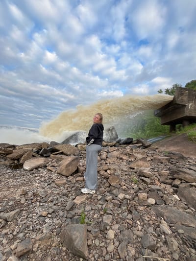 A young blonde woman stands on a rocky shoreline, looking over her left shoulder at the camera. She is wearing a black hooded sweatshirt with white stripes on the sleeves and gray sweatpants. Behind her, a large plume of murky water is being forcefully discharged from a concrete structure. The sky is filled with dramatic, cloudy formations. The surrounding area is covered in various sizes of rocks and pebbles, with some sparse green vegetation visible on the right side of the frame. The location is identified as Chernushka, Russia, though specific landmarks are not discernible in the image to pinpoint an exact spot within the city. The overall scene suggests a potentially industrial or infrastructural water release in an outdoor, somewhat rugged environment.