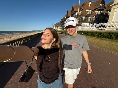 A selfie shows two people walking outdoors. A man wearing a gray t-shirt and white hat with a logo walks alongside a woman in a black shirt and jeans. The background includes a brick walkway, a building with a Tudor-style facade, a green strip of grass, a beach, and the sky. The image appears to be taken sideways, with the sky at the bottom and the building at the top.
