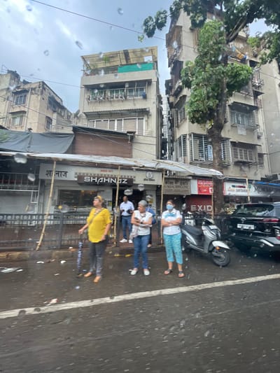 The image shows a street scene in Mumbai, India, on a rainy day. Several people are standing on the sidewalk, some are looking at their phones, and others are looking in different directions. There are shops and buildings in the background. A scooter and a black SUV are parked on the street. The overall atmosphere is wet and overcast, with visible raindrops on the camera lens.
