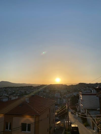 This is a vertical, eye-level shot of a sunset over a hilly city, likely Sandanski, Bulgaria. The sun, a bright, warm orb, sits low on the horizon, casting a golden glow across the sky and the dense urban landscape below. The sky transitions from a soft, clear blue on the left to a hazy, pale yellow near the sun, indicating a clear day with the sun setting.
The scene is dominated by the sprawling city, which appears to be built on hills, with buildings of varying heights and architectural styles densely packed together. Many buildings have red-tiled roofs, which are illuminated by the setting sun. Power lines crisscross the scene, adding a layer of urban detail. A road with a dark-colored car can be seen on the right side of the frame. In the foreground, a building with a prominent red-tiled roof and windows is visible. The overall mood is serene and peaceful, typical of a sunset. The time of day is clearly sunset due to the position of the sun and the warm, low-angled light. No people are clearly visible, but the presence of cars and buildings suggests an inhabited area. There is no visible text in the image.