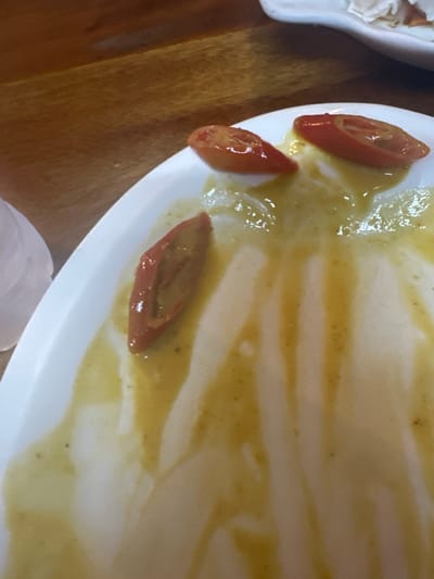 The image displays a close-up, high-angle view of a partially consumed meal on a white, oval-shaped plate, set within a dining environment in Tambon Ko Tao, Thailand. The plate is largely covered with streaks of viscous, yellowish-green sauce, which contains small dark specks. Three elongated, longitudinally sliced red-orange pepper pieces, each approximately 3-4 cm long, rest on the plate amidst the sauce remnants. The background reveals a warm-toned wooden table surface with visible grain. A blurred white object, possibly a glass or another dish, is partially visible on the far left edge, and another out-of-focus white object, likely a plate, is present in the upper right. No individuals are depicted in the scene.