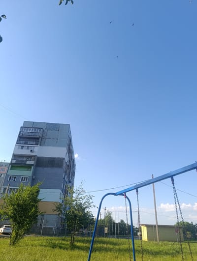 A low-angle shot captures a bright, clear blue sky with a few wispy clouds. In the foreground, a blue metal swing set with heavy chains is visible, suggesting a playground setting. To the left, a tall, multi-story apartment building with a somewhat weathered facade stands against the sky, featuring balconies and windows. Several trees with lush green foliage are interspersed between the building and the playground. A white car is parked on the left side of the frame. The ground is covered in green grass, indicative of a sunny day. In the distance, a small yellow building with a red sign is visible, along with other trees and structures, hinting at an urban or suburban environment. Several small birds are silhouetted against the sky, suggesting they are in flight. The overall impression is one of a pleasant, ordinary day in an outdoor public space. No discernible text is visible in the image.