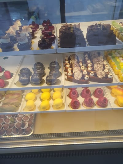 The image displays a refrigerated display case filled with an assortment of pastries and chocolates, likely in a bakery or patisserie in Fergana City, Uzbekistan. The display case is divided into multiple shelves, showcasing a variety of meticulously crafted desserts.
On the top shelf, there are several dark chocolate cakes topped with decorative elements, including what appear to be chocolate shavings and gold leaf. Next to these, to the right, are rows of small, bright yellow pastries, some adorned with green accents. To the left of the chocolate cakes are smaller, dark-colored individual desserts topped with berries and a swirl of cream, some with an accompanying decorative element resembling a fan.
The middle shelf features rows of round, dark purple desserts, with three in the front row. To their left, on the far left of this shelf, are two heart-shaped pink pastries. To the right of the purple desserts, there is a tray of elongated, pale-colored pastries, some with a pinkish hue, and further to the right, several yellow, lemon-shaped pastries are arranged on gold bases.
The bottom shelf contains a tray filled with numerous small, individually wrapped chocolates and what appear to be chocolate-covered strawberries. Some of the chocolates are nestled in white paper liners. To the right of the chocolates, there are two mango-shaped pastries on gold bases, and to the right of those, there are additional yellow, lemon-shaped pastries.
The lighting in the display case highlights the glossiness of the desserts. The overall environment suggests a commercial setting focused on high-end confectionery. There are no visible people in the image.
