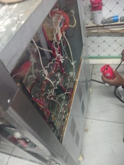 An open electrical panel reveals a complex network of wires and components. Red, white, and other colored wires are intertwined within the metal enclosure. A person holds a red electrical connector nearby, seemingly inspecting or working on the equipment. The background includes white-tiled walls and additional electrical fixtures, suggesting an industrial or commercial setting.