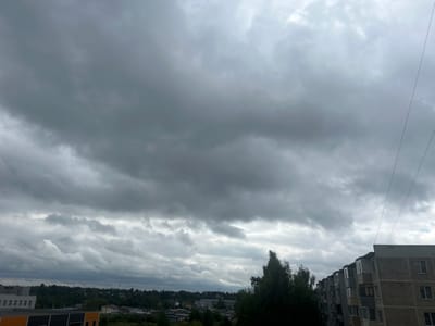 The image depicts a cloudy sky over a city. In the foreground, a modern building with multiple windows and balconies dominates the right side of the frame. To the left, a lower-rise building with orange and black accents is visible. The middle ground is filled with trees and other smaller buildings, suggesting a residential or industrial area. The background features a dense line of trees and the overcast sky, characterized by dark, heavy clouds. The scene appears to be an urban or suburban landscape in Kokhma, Russia. There are no people or discernible actions taking place in the image.