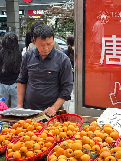 A man in a navy shirt is selling baskets of loquats at a street market in Nan'an District, China. The fruit is displayed in red plastic baskets. The street scene includes other pedestrians, street signs in Chinese, and a food or product advertisement in the background.