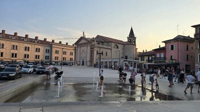 This is a medium shot of a public square in Peschiera del Garda, Italy, at dusk. A large, light-stone building with many arched windows dominates the left side of the frame, suggesting it might be a historical or municipal building. To its right stands a large, tan church with a prominent bell tower. In front of the church, there are several outdoor dining areas with tables and umbrellas, and people are seated, enjoying meals or drinks.

The foreground features a shallow water fountain where several children are playing, splashing in the water with visible joy and excitement. Adults are also present, some watching the children, others socializing. A variety of cars are parked in the background on a street behind the buildings on the left, and there is a construction crane visible above the buildings. The sky is a soft blue with hints of orange, indicating it is either sunrise or sunset. The overall atmosphere appears relaxed and pleasant, with people enjoying a warm evening outdoors in a lively town square. The architecture and the presence of a church strongly suggest a European setting, consistent with the provided location context.
