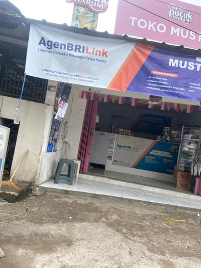 The image shows the storefront of a small shop in Baleendah, Indonesia. The shop is located at coordinates -6.996 latitude and 107.631 longitude. The storefront is adorned with several signs, including one that reads "AgenBRILink Layanan Transaksi Keuangan Tanpa Kantor" and another that reads "Toko Musti." The shop offers various financial services, including cash withdrawals, deposits, and bill payments. The shop is open and there is a counter with a sign that reads "AgenBRILink" above it. There is also a broom and a bucket of water outside the shop. The shop appears to be open for business.