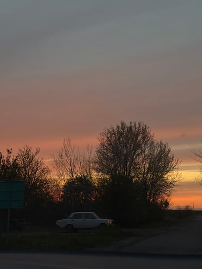 The image, taken in Alexandrovka, Russia, captures a serene moment at either sunset or sunrise. The sky dominates the upper and left parts of the frame, displaying a striking gradient of colors. It transitions from a soft, muted grey-blue on the far left to warmer hues of pink, vibrant orange, and bright yellow on the right, indicating the low angle of the sun.
Dense clusters of trees, primarily consisting of bare, intricate branches, are silhouetted against this colorful sky, occupying the right side of the composition. A dark road or unpaved path runs horizontally along the bottom of the image.
Parked on the right side of this path is a light-colored sedan, possibly white or light grey. Its rear section is visible, and one of its taillights appears to be faintly illuminated. Partially obscured by the foreground trees, a dark green rectangular road sign is visible in the bottom right corner, though no text or symbols can be discerned.
No people or explicit activities are visible; the scene conveys a sense of quietude and natural beauty. The clear sky and vivid atmospheric colors are the main focus, suggesting fair weather. The overall impression is one of a peaceful, contemplative landscape at the transition between day and night.
