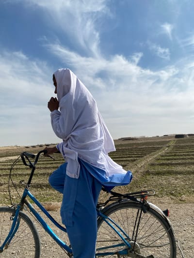 A person is depicted riding a blue bicycle on a dirt path in Nakhonay, Afghanistan. The individual, dressed in a white head covering that flows over a blue garment, is seen in profile, facing left, with hands positioned near the mouth. The bicycle has a rear rack and appears aged. To the right of the path, a cultivated field with distinct rows extends into the midground. The background consists of a flat, arid landscape under a blue sky with scattered white cirrus clouds, with small, low-lying structures visible in the distance on the horizon.