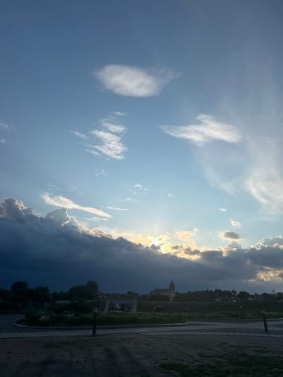A vertical, wide-angle photograph captures a dramatic sky over a semi-urban landscape. The upper half of the image is dominated by a bright blue sky filled with various cloud formations. A few wispy cirrus clouds are scattered high up, while more substantial cumulus clouds drift lower. The sun is partially obscured by a large, dark bank of clouds on the left, with rays of golden light breaking through on the right side of this cloud bank.
The lower half of the image depicts a town or village silhouetted against the sky. A prominent, red-roofed church with a tall steeple is visible in the distance, slightly right of center. A line of trees forms a dark green band between the clouds and the buildings. In the foreground, there is a paved area with a sandy or gravelly surface, marked with dark gray square tiles. A curved, landscaped area with green bushes, some flowers, and bollard-style lights is situated between the foreground pavement and the silhouetted town. The overall impression is of a scenic view, possibly during sunset or sunrise, with a storm approaching or receding. The location is identified as Zhukhavichy Sel'ski Savet, Belarus.