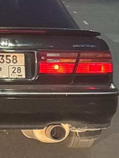 A rear three-quarter view shows the back of a dark-colored sedan. The license plate is visible on the left side of the trunk, displaying "358" above "28" and "RUS" with the Russian flag emblem below. To the right of the license plate are the red taillights of the vehicle, which appear to be illuminated. Above the taillights, the word "INSPIRE" is subtly visible on the trunk lid. A chrome exhaust pipe is visible underneath the rear bumper on the right side. The vehicle is parked on a paved surface, possibly a street or parking lot, with some white lines visible in the background suggesting a road. The lighting conditions indicate it may be dusk or nighttime. The overall scene is consistent with an urban environment in Blagoveshchensk, Russia.