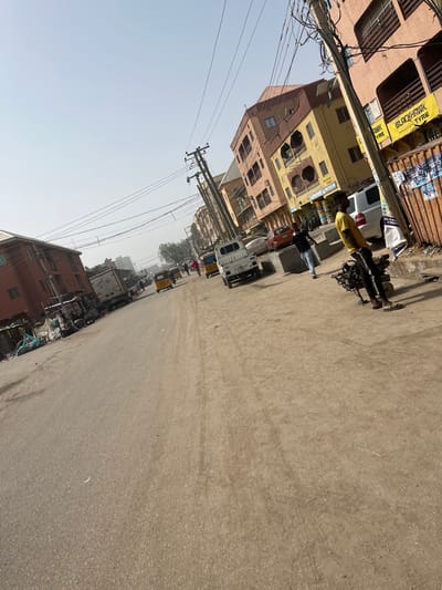 This image depicts a street scene in Sabon Gari West, Nigeria, under clear, hazy daylight. The paved road, exhibiting tire tracks and minor dirt accumulation, occupies the lower and central portions of the frame, leading into the distance. Multi-story commercial and residential buildings with varying facades (tan, yellow, orange-brown) line both sides of the street. Utility poles, heavily laden with multiple electrical and communication wires, are prominent on the right side and extend along the road.

On the immediate right, a male individual wearing a yellow short-sleeved shirt, dark track pants, and sandals stands, actively pushing or operating a dark-colored, two-wheeled piece of machinery, possibly a generator or pump. Behind him, a "BLACKHAWK TYRE" sign is visible on a building, alongside a "B. ROYALE BICYCLE" sign further down. A white pickup truck is parked on the right, and an orange car is visible behind it. Another male in a dark shirt and pants walks across the street towards the center of the frame. In the mid-ground, multiple yellow tricycle taxis (keke napep) and other vehicles are present on the road. The left side of the street features more buildings and accumulated debris or merchandise near the roadside. Several other distant figures are discernible moving along the street. The sky is a pale, uniform blue, indicating an overcast or hazy atmospheric condition.
