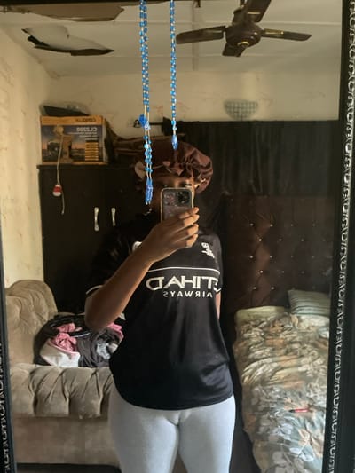 This image depicts an indoor scene in Ajaokuta, Nigeria, captured as a mirror reflection. A single individual, presumably female, is centered in the frame, taking a selfie with a smartphone held to obscure her face. She wears a brown satin sleep bonnet, a black short-sleeved t-shirt with the reversed "ETIHAD AIRWAYS" logo in white, and light grey ribbed leggings.
The background, visible in the mirror, shows a room with a damaged ceiling featuring peeled plaster and paint, revealing a darker surface underneath. A brown three-bladed ceiling fan is mounted. Two strands of blue beaded decorations hang vertically in front of the mirror.
To the left, a dark brown or black two-door wardrobe with silver handles is visible. On top of the wardrobe, a brown cardboard box labeled "CROWN" and "CL 3500" (reversed) is present, along with other indeterminate objects. A small white and red object, possibly a decoration, hangs near the wardrobe. Below the wardrobe, a light-colored, tufted armchair or sofa is partially visible, with a pile of folded clothes in various colors (pink, black, white) on its seat.
To the right, a dark brown tufted headboard with light-colored button details is against the wall. Below the headboard, a bed is made with a patterned duvet or blanket in shades of green, beige, and white. A small red object, possibly a marker or pen, rests on the bed. A small, round, white decorative object or light fixture is reflected on the upper right wall. The mirror itself has a dark, ornate frame.