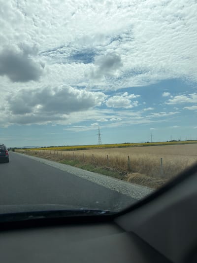 The image captures a view from inside a vehicle driving on a paved road. To the left of the road, a dark-colored car is visible. The road is bordered by a gravel shoulder and then a field of dry, tall grass with sparse wooden posts. Beyond the dry field, a vibrant yellow field of sunflowers stretches across the landscape. In the distance, several tall power pylons are scattered across the horizon. The sky is filled with a mix of white and gray clouds, with patches of blue sky visible. The overall scene suggests a rural setting in Bulgaria, possibly in the vicinity of Загорци, given the agricultural landscape.