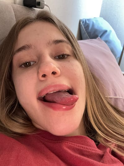 A close-up, slightly angled selfie shows a young woman lying down, wearing a red shirt and looking at the camera. Her eyes are brown, and her hair is long and blonde. She has her tongue sticking out and her lips are glossy. The background shows pillows and a wall. The image was taken in Vsevolozhsk, Russia.