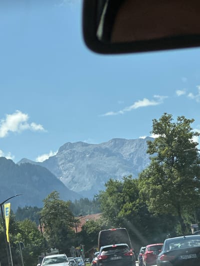The image captures a mountainous landscape behind a street scene in Berchtesgaden, Germany. In the foreground, several cars are visible on a road, suggesting traffic congestion. A yellow flag with a blue "E" logo is planted on the left side of the road, likely indicating a store. Green trees and some buildings with red roofs are interspersed along the street. In the background, a vast mountain range dominates the horizon, characterized by rocky slopes and sparse vegetation. The sky is clear blue with a few scattered clouds. The overall impression is of a scenic drive through a town nestled in a mountainous region.