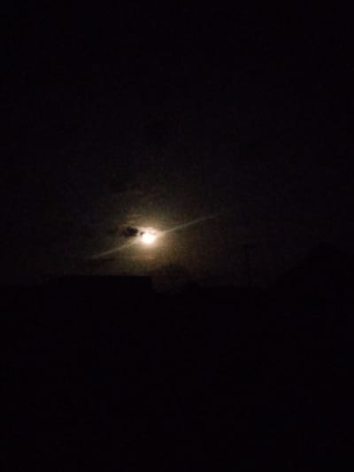 The image depicts a dimly lit outdoor scene, likely at night. The most prominent feature is a bright, circular object in the upper-middle section, which appears to be the moon, partially obscured by clouds. A faint beam of light extends from the moon diagonally to the left. In the lower portion of the image, dark silhouettes of what seem to be buildings or structures are visible. The overall environment is dark and atmospheric, suggesting a night sky. The provided context indicates this scene is located in Mangu Town, Nigeria.